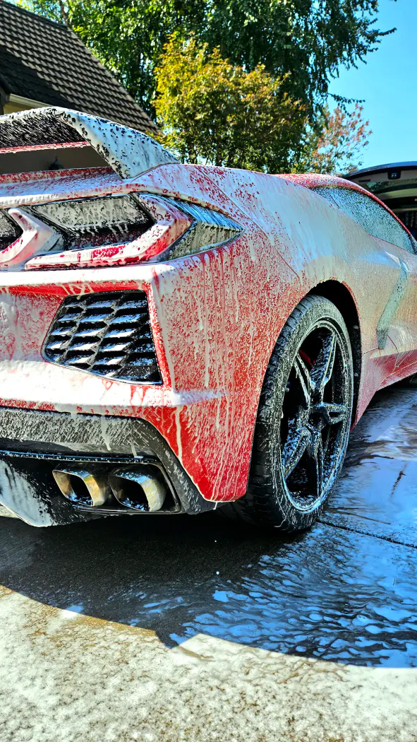 Red sports car being washed, covered in white soap suds, with foam dripping down its sides, parked outdoors under a clear sky.