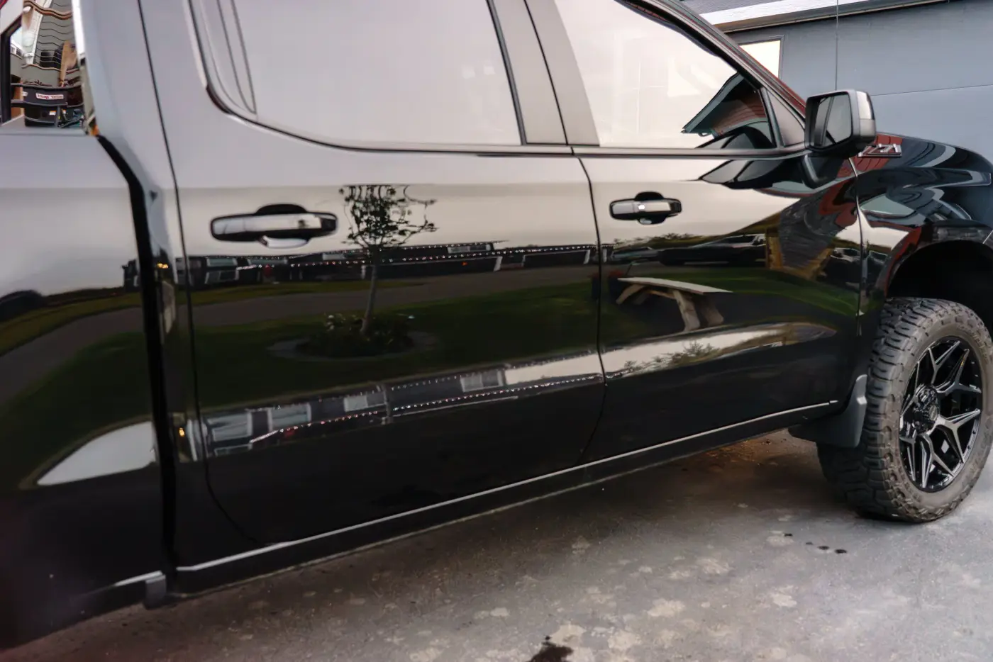 Close-up of a black pickup truck parked outside, showing the side doors, side mirror, and front wheel with off-road tires, reflecting trees and surroundings on its shiny surface.