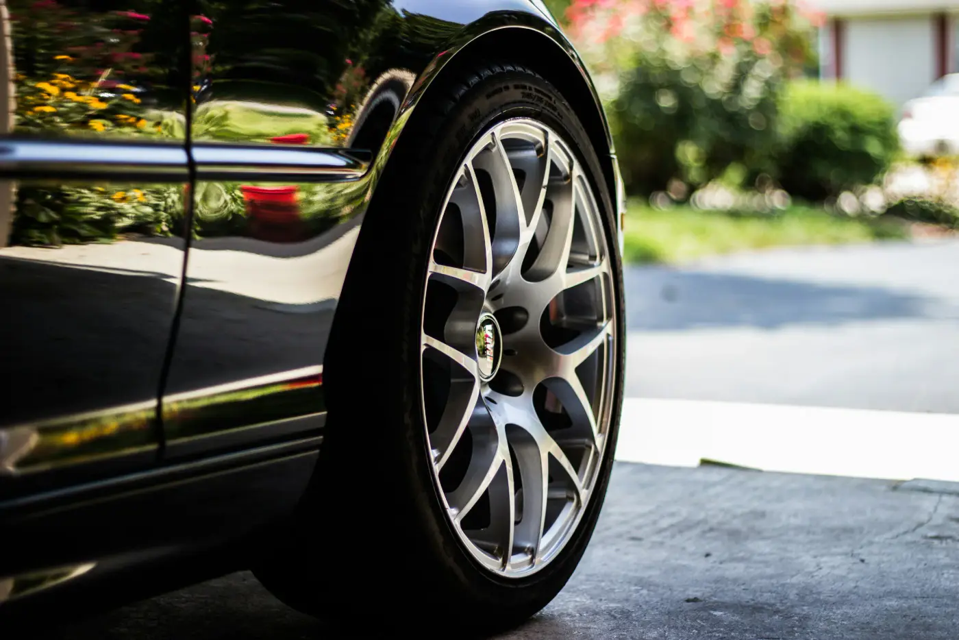Close-up of a car wheel with alloy rims and reflective paint, parked in a driveway with garden in the background.