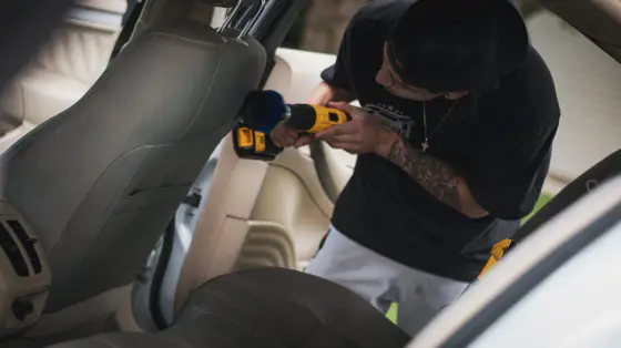 A person with tattoos, wearing a black cap and black t-shirt, using a yellow cordless drill to clean or repair the inside of a vehicle.